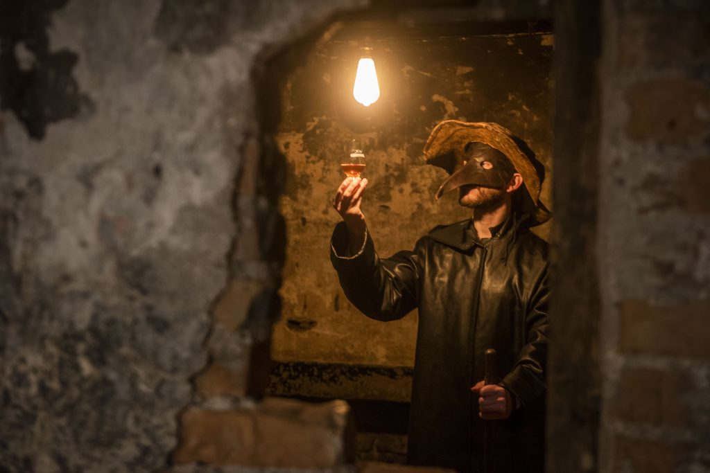 Tour guide holding a dram of whisky while wearing a plague doctor mask. 