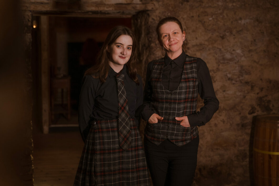 Two Guest Services team members from The Real Mary King's Close. One wears a kilt and the other a waistcoat embroidered with a Kinloch Anderson tartan. 