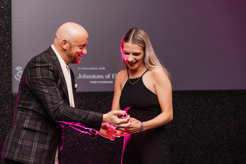 Katie Scott (pictured right, black dress, blonde hair), receives Scottish Thistle Award 2023 from man in tartan suit 